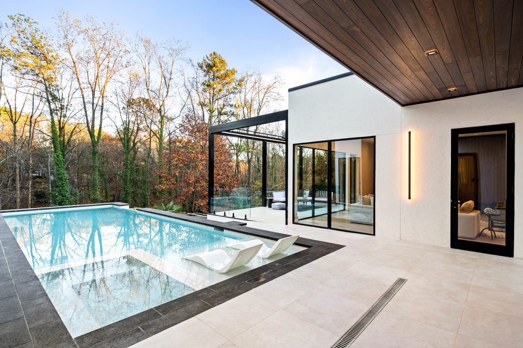 Modern minimalist pool with white lounge chairs submerged on a sun shelf, next to a light-tiled terrace and a contemporary house with large glass windows.
