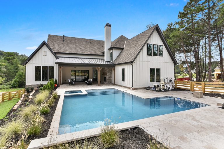 Rectangular pool with an integrated spa in front of a white modern farmhouse with dark roofs, a wooden-post porch, and a light stone deck.