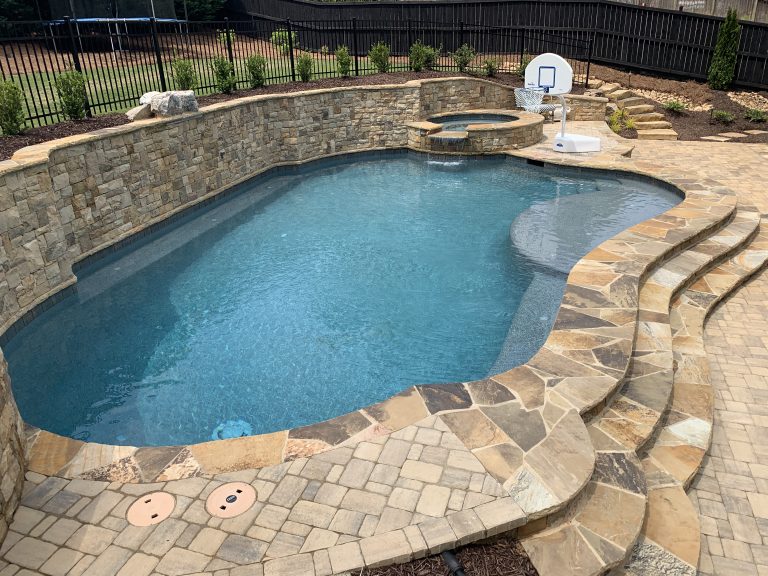 Freeform pool with natural stone coping, an elevated jacuzzi with a waterfall, and a rustic stacked stone retaining wall in a paved backyard.