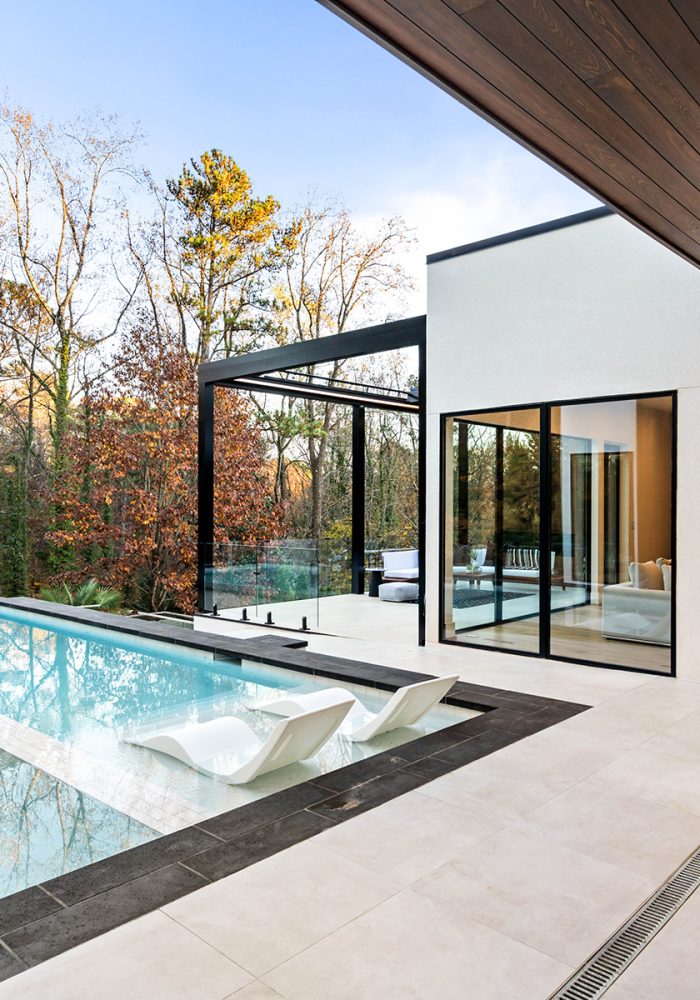 Modern minimalist pool with white lounge chairs submerged on a sun shelf, next to a light-tiled terrace and a contemporary house with large glass windows.