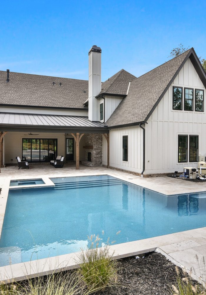 Rectangular pool with an integrated spa in front of a white modern farmhouse with dark roofs, a wooden-post porch, and a light stone deck.