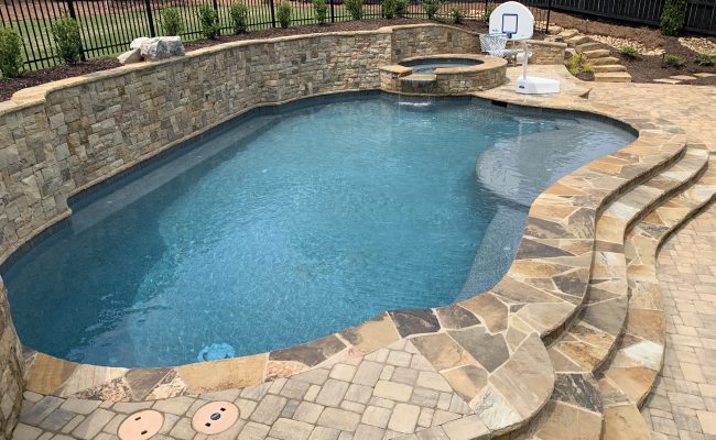 Freeform pool with natural stone coping, an elevated jacuzzi with a waterfall, and a rustic stacked stone retaining wall in a paved backyard.