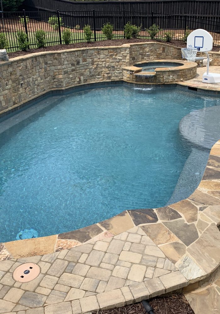 Freeform pool with natural stone coping, an elevated jacuzzi with a waterfall, and a rustic stacked stone retaining wall in a paved backyard.